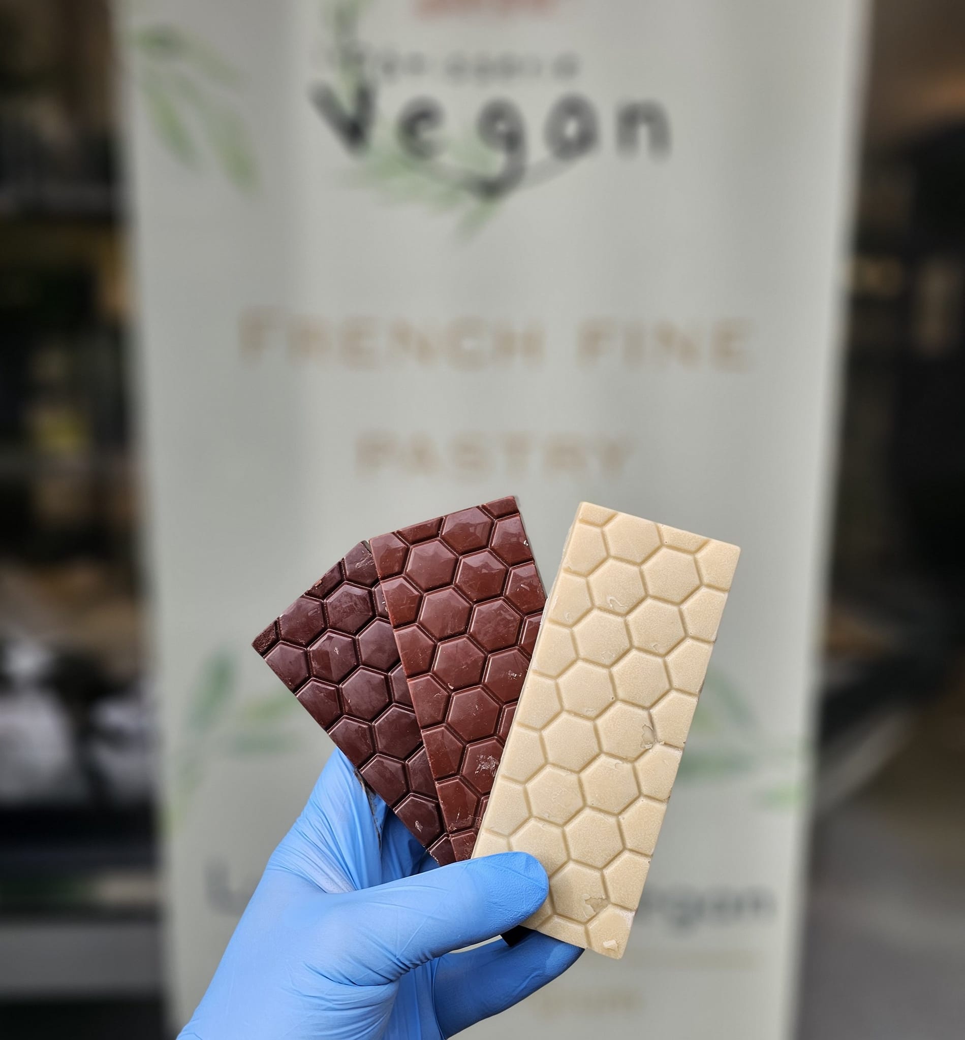 Devant la devanture de La Pâtisserie Vegan à Liège, trois délicieux chocolats végans se présentent en une irrésistible symphonie de saveurs et de couleurs :

Chocolat Noir :
Une tablette d’un noir profond, luisante et cassante.
Des notes intenses de cacao, légèrement acidulées.
Mots-clés : Noir, cacao, gourmandise, végétalien.
Chocolat Blanc :
Une douceur immaculée, crémeuse et fondante.
Des arômes de vanille et de beurre de cacao.
Mots-clés : Blanc, vanille, onctuosité, pâtisserie végane.
Chocolat au Lait :
Une tablette veloutée, couleur caramel, avec une texture soyeuse.
Des saveurs douces et réconfortantes, rappelant l’enfance.
Mots-clés : Lait, caramel, douceur, gourmandise.
Ces chocolats, confectionnés avec passion par Nicolas Bastille, sont la quintessence de l’art pâtissier végétal. Ils invitent à une dégustation exquise, tout en respectant nos convictions et notre planète. 🌱🍫